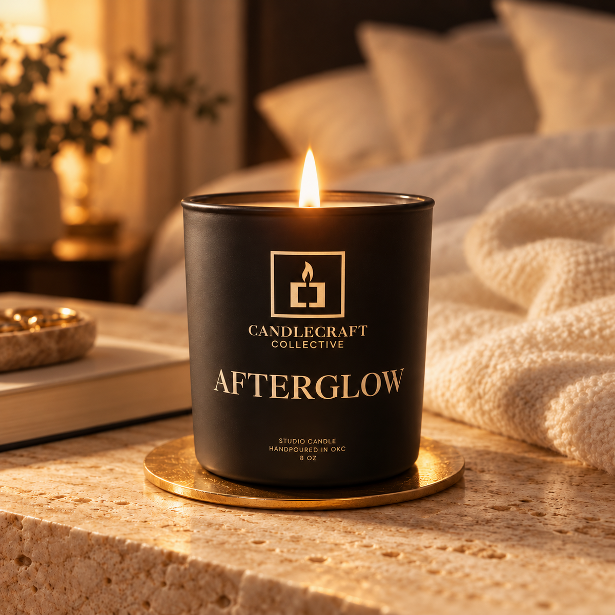 A black candle jar with a gold logo and the text 'CANDLECRAFT COLLECTIVE AFTERGLOW' sits on a gold coaster on a stone surface, with a softly lit bedroom scene in the background.